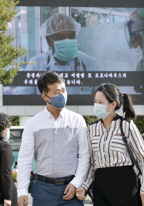북한 평양역 앞을 마스크를 쓴 남녀가 걸어가고 있다. 이들의 뒤에 있는 대형 모니터에는 세계 코로나19 감염 상황을 전하는 영상이 비치고 있다.〈사진=연합뉴스〉