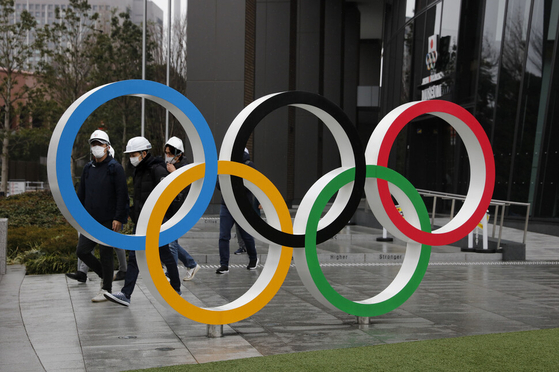 지난해 3월 일본 도쿄 국립경기장 주변에 설치된 올림픽 로고 뒤를 마스크를 쓴 시민들이 지나가고 있다. 〈사진=AP 연합뉴스〉